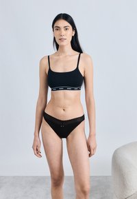 Braletă neagră cu bretele subțiri și bandă cu logo, combinată cu chiloți negri cu dantelă și detaliu cu fundiță mică. Textură netedă, design conținut.