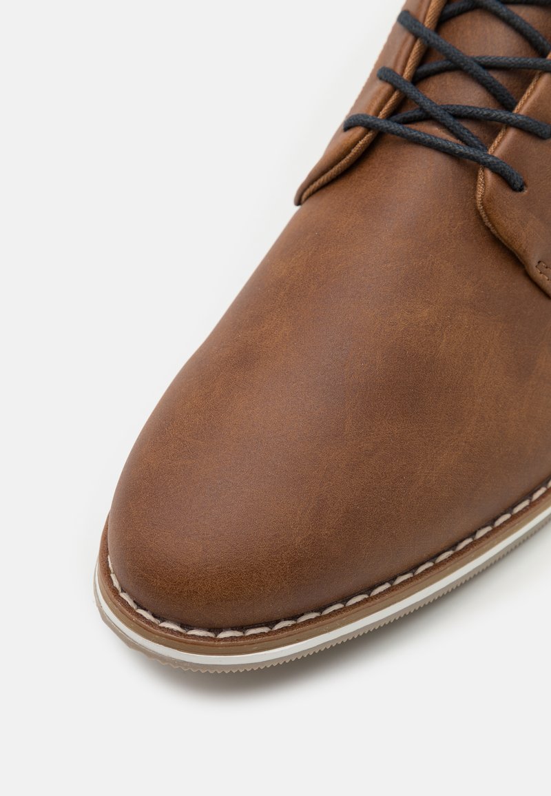 Zapato de cuero marrón con una textura suave, punta redondeada y cordones negros. Presenta costuras blancas a lo largo del borde de la suela.