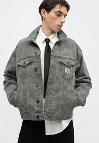 Chaqueta de mezclilla gris con patrón de camuflaje, dos bolsillos en el pecho, cierres de botones y un parche del logo de Carhartt en el lado izquierdo.