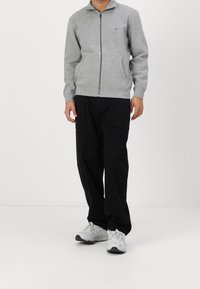 Grau melierte Reißverschlussjacke mit hohem Kragen und zwei Fronttaschen. Kombiniert mit weit geschnittenen schwarzen Hosen und weißen Turnschuhen.