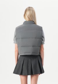 Femme blonde portant un pull gris à manches courtes, un gilet matelassé gris et une jupe plissée gris foncé, debout de dos sur un fond blanc.