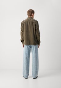 Olive green corduroy jacket with a shirt collar, paired with light blue baggy denim jeans. Viewed from the back against a neutral background.
