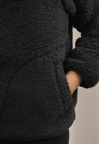 Pull-over en polaire noire avec une surface texturée et duveteuse. Comprend une poche avant et des manches avec poignets pour la chaleur et le confort.