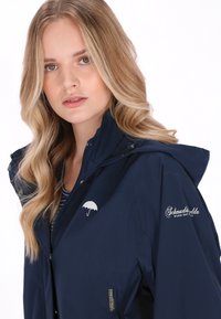 Granatowa wodoodporna kurtka z kapturem, z białym logo parasola, zamkiem błyskawicznym oraz haftowanym nadrukiem na rękawie i klatce piersiowej.