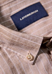 Ljusbrun linneskjorta med tunna vita ränder, med en marinblå etikett märkt "LINDBERGH" och en rund knapp vid kragen.