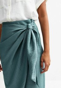 A teal wrap skirt made of textured fabric, featuring a tied knot detail at the waist and a soft drape extending down the side.