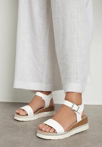 Białe sandały na platformie mają szeroki pasek z przodu, zapięcie na kostce oraz teksturowany obcas w kształcie klinu z błyszczącym akcentem wzdłuż podeszwy.