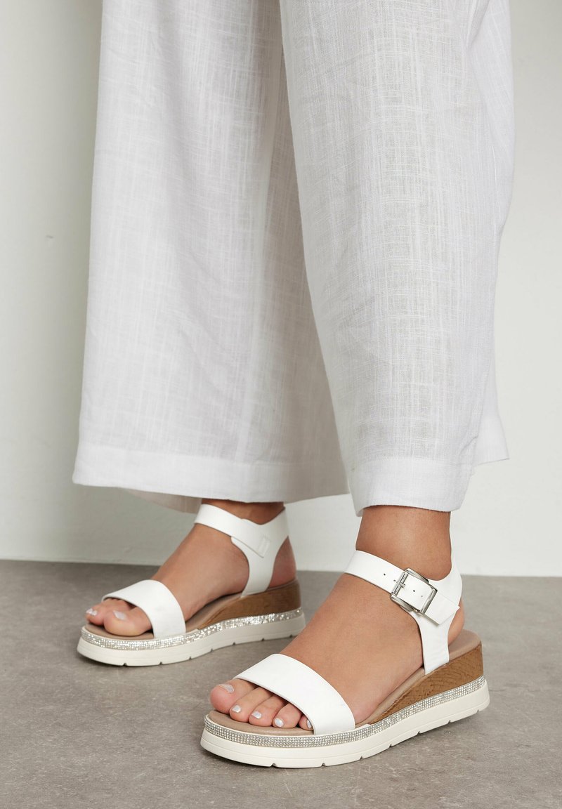 Białe sandały na platformie mają szeroki pasek z przodu, zapięcie na kostce oraz teksturowany obcas w kształcie klinu z błyszczącym akcentem wzdłuż podeszwy.
