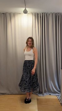 Γυναίκα με άσπρο αμάνικο μπλουζάκι και μακρύ μαύρο φούστα με σχέδια στέκεται σε ξύλινο πάτωμα μπροστά από γκρι κουρτίνες κάτω από φως οροφής.