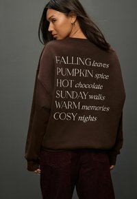 Barna kapucnis pulóver fehér felirattal a hátán: "HULLÓ levelek, TÖK fűszer, FORRÓ csokoládé, VASÁRNAPI séták, MELEG emlékek, KÉNYELMES éjszakák."