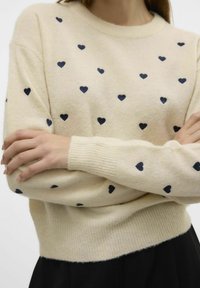 Pull beige avec des motifs de cœurs navy, col rond et poignets côtelés ; fabriqué en matériau tricoté doux, coupe légèrement décontractée.