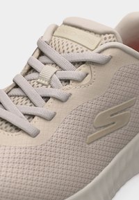 Μπεζ αθλητικό παπούτσι Skechers με ύφασμα από δίχτυ, παχιά λευκά κορδόνια, επενδεδυμένο γιακά και υφασμένο πέλμα, εμφανιζόμενο σε κοντινή γωνία.