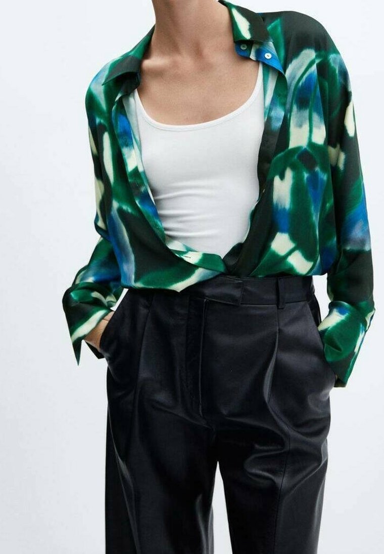 Persona che indossa una camicia a maniche corte a bottone con fantasia verde, blu e bianca sopra una canottiera bianca, abbinata a pantaloni neri a vita alta, con le mani in tasca.