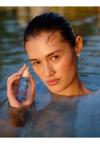 Blå glasflaske med Lumene Nordic Hydro Aqua Hydration Serum med en hvid pipettehætte, holdt af en person med vådt hår, delvist nedsænket i vand.