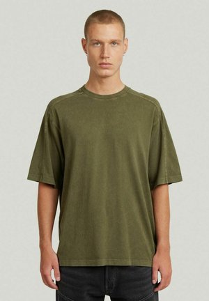 Giovane uomo con capelli corti che indossa una maglietta oversize verde oliva e pantaloni neri, in piedi davanti a uno sfondo chiaro e uniforme.