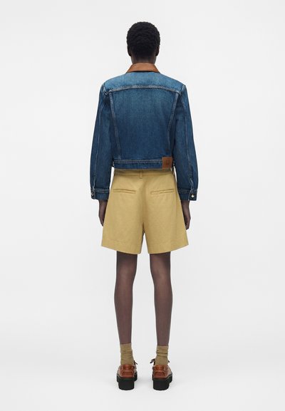 Άτομο που φοράει μπλε τζιν μπουφάν με καφέ γιακά, μπεζ σορτς με τσέπες πίσω, ταμπ καλτσάκια και καφέ παπούτσια με πλατφόρμα, στέκεται με την πλάτη γυρισμένη.