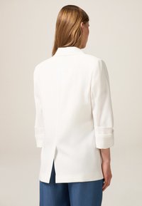 Blazer blanco con un corte entallado, mangas de 3/4 enrolladas, detalle de abertura en la parte posterior y textura de tela suave, combinado con pantalones azules.