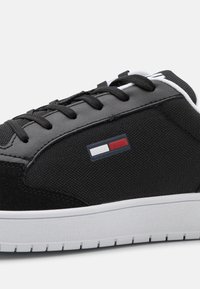 Baskets noires et grises avec lacets noirs, panneaux en tissu texturé, semelle blanche et un petit logo rectangulaire rouge, blanc et bleu sur le côté.