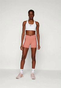 Balta sporta krūšturis ar Nike logo, persiku krāsas šorti un gaiši rozā sporta apavi. Apģērbs ir ar pieguļošu dizainu un gludu tekstūru.