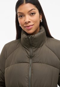 Giacca a vento imbottita verde oliva con collo alto, chiusura a zip e design trapuntato. Tessuto liscio con un aspetto leggero e una lucentezza sottile.