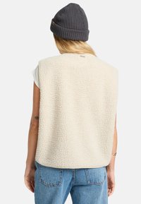 Fleece vest in cream with a textured surface, sleeveless design, and a rounded hem. Paired with a gray knitted beanie.