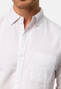 Chemise boutonnée blanche en coton avec un col, dotée d'une poche poitrine unique et d'un tissu texturé. Les boutons sont blancs et au design classique.
