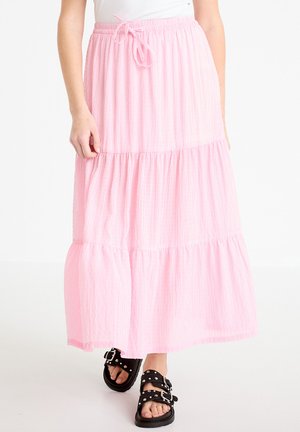 Vrouw draagt een lange, gelaagde roze rok met een trekkoord in de taille en zwarte sandalen met studs en gespen.