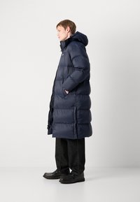 Blå pufferjakke med hette, quiltet design, sidelommer og glidelås. Brukt over mørke denimbukser og svarte sko.