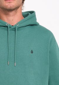 Sudadera verde menta con un bolsillo tipo canguro, cordones ajustables y un pequeño logo negro en el pecho izquierdo. Tejido suave y texturizado.