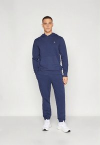 Felpa con cappuccio blu navy e pantaloni jogger abbinati in tessuto morbido, con tasca a marsupio e dettaglio logo sottile, abbinati a sneakers bianche.