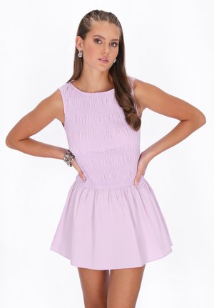 Kvinde med langt brunt hår iført en ærmeløs lavendelfarvet kjole, stående med hænderne på hofterne foran en hvid baggrund.