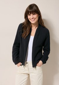 Zwarte zip-up jacket van een gestructureerde stof, met voorzakken en elastische manchetten, gedragen over een witte top en lichte broek.