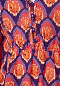 Tissu à motif floral violet et orange avec boutons violets et couture froncée à la taille sur un vêtement.