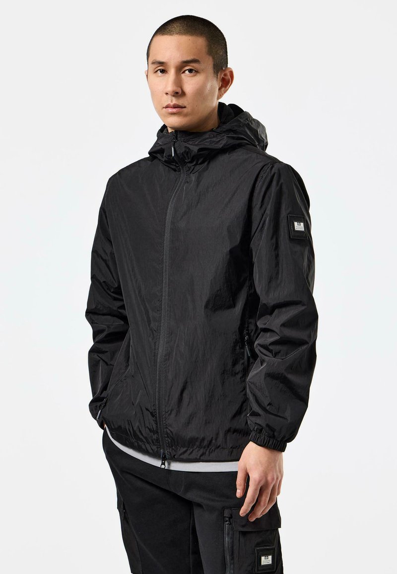 Schwarze leichte Jacke mit Kapuze, durchgehendem Reißverschluss, elastischen Bündchen, Seitentaschen und einem kleinen Patch am linken Ärmel.
