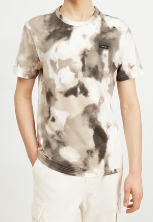 Man draagt beige en bruine tie-dye korte mouw T-shirt met Calvin Klein-logo op de borst, gecombineerd met witte cargobroek.