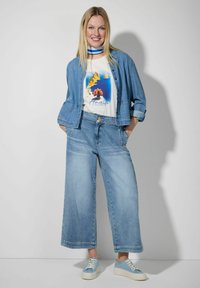 Hellblaue Jeansjacke, weißes T-Shirt mit grafischem Design und blauen Akzenten, weit geschnittene, verwaschene Jeans und hellblaue Sneakers. Weiche Textur, lockere Passform.