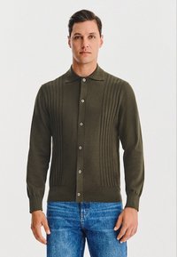 Oliwkowy kardigan z pionowym prążkowanym wzorem, z kołnierzykiem i zapięciem na pięć guzików, zestawiony z niebieskimi dżinsami.