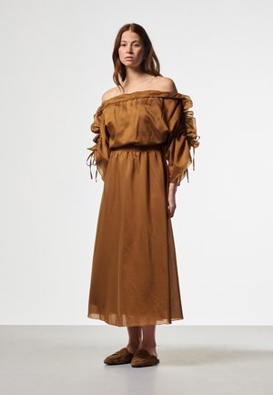 Frau mit langen braunen Haaren, die ein weites, schulterfreies braunes Kleid mit gerafften Ärmeln und braunen Slippern trägt, steht vor einem schlichten Hintergrund.