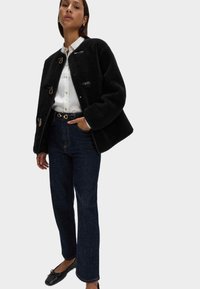 Giacca in pile nera con chiusura a bottoni a pressione, abbinata a una camicia bianca con bottoni e jeans scuri a vita alta. Ballerine nere completano l'outfit.