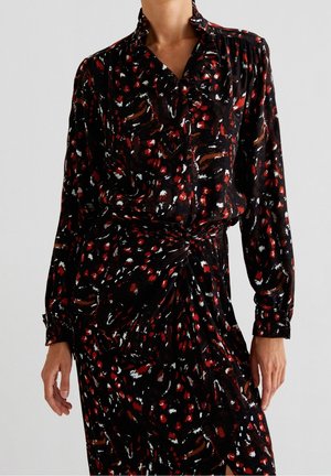 Femme portant une robe noire à manches longues avec des taches abstraites rouges, blanches et brunes, avec une taille froncée et un col.