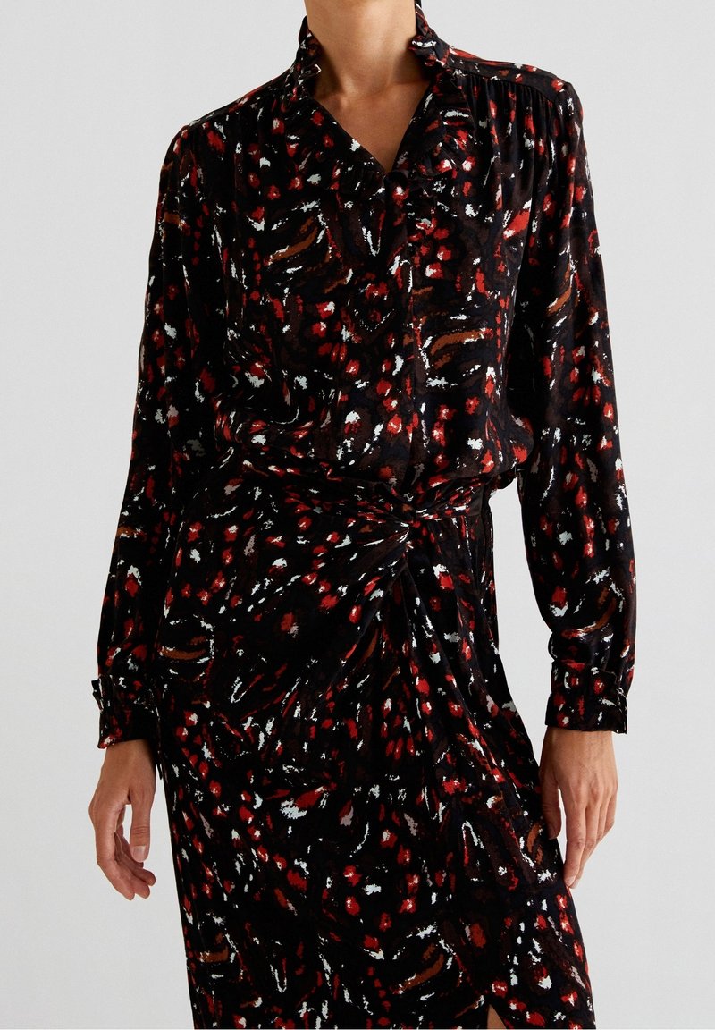 Femme portant une robe noire à manches longues avec des taches abstraites rouges, blanches et brunes, avec une taille froncée et un col.
