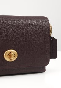 Tummanpunainen nahkainen olkalaukku, jossa on kupruileva pinta, kultaiset metalliosat, magneettinen suljin ja COACH-logo painettuna tunnisteessa.