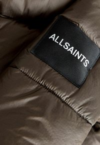 Brun pufferjacka i tyg med en slät, glänsande yta. Har en svart lapp med "ALLSAINTS" broderat i vita bokstäver.