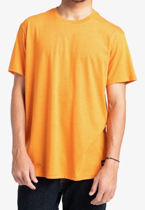 Mann trägt ein schlichtes, knalloranges T-Shirt mit kurzen Ärmeln und schwarze Hose, steht mit entspannten Armen seitlich am Körper vor weißem Hintergrund.