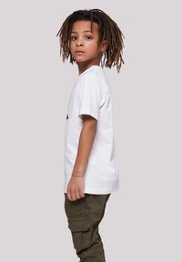 Weißes Baumwoll-T-Shirt mit kurzen Ärmeln, das ein rotes Design zeigt. Olivgrüne Cargo-Hose mit Taschen. Kind mit Dreadlocks, das seitlich schaut.