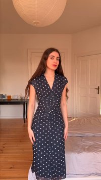 Γυναίκα με μακριά καστανά μαλλιά, που φοράει ένα μαύρο φόρεμα με λευκές πουά, στέκεται σε ένα υπνοδωμάτιο με ξύλινο πάτωμα και στρογγυλό φωτιστικό οροφής.