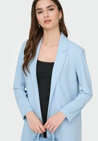 Gaiši zils žakete ar piegriezumu, garām piedurknēm un atloku apkakli, kopā ar melnu topu un rokā turot mazu zilu somu.