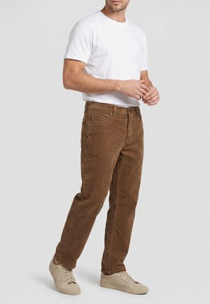 Hombre con camiseta blanca lisa de manga corta, pantalones de pana marrones y zapatillas beige claras, de pie con las manos entrelazadas.