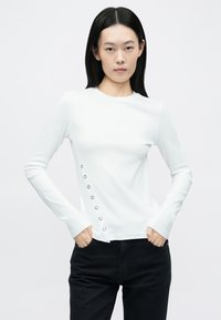 Biely dlhý rukáv s priliehavým strihom, ktorý obsahuje rad čiernych gombíkov s otvormi pozdĺž ľavej strany. Hladká textúra a okrúhly výstrih.