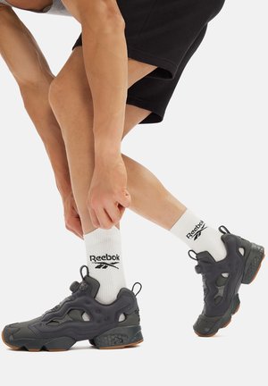 Czarne sneakersy Reebok z teksturowaną górą, widoczną amortyzacją i brązową gumową podeszwą, w połączeniu z białymi skarpetkami z detalem logo.